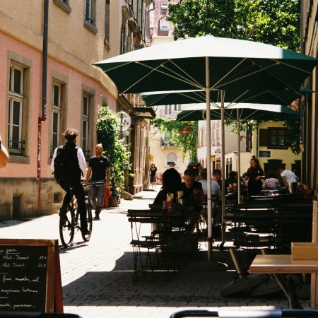 A street in Europe with a man on a bike and patrons at a cafe.