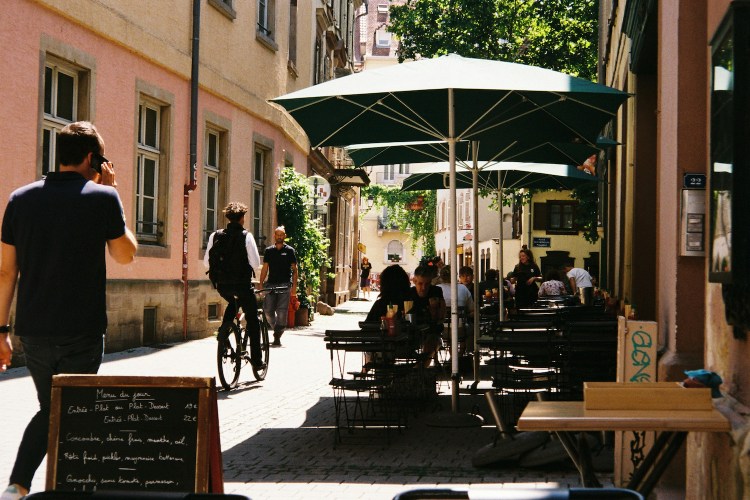 A street in Europe with a man on a bike and patrons at a cafe.