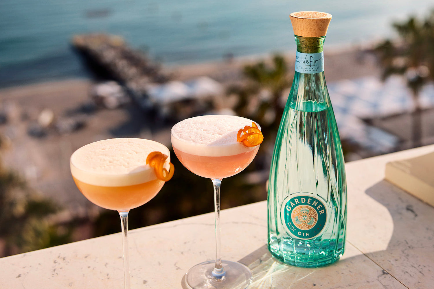 two cocktails next to a bottle of The Gardener Gin on a ocean background