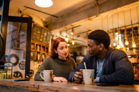 two people sitting and talking next to each other with coffee mugs.