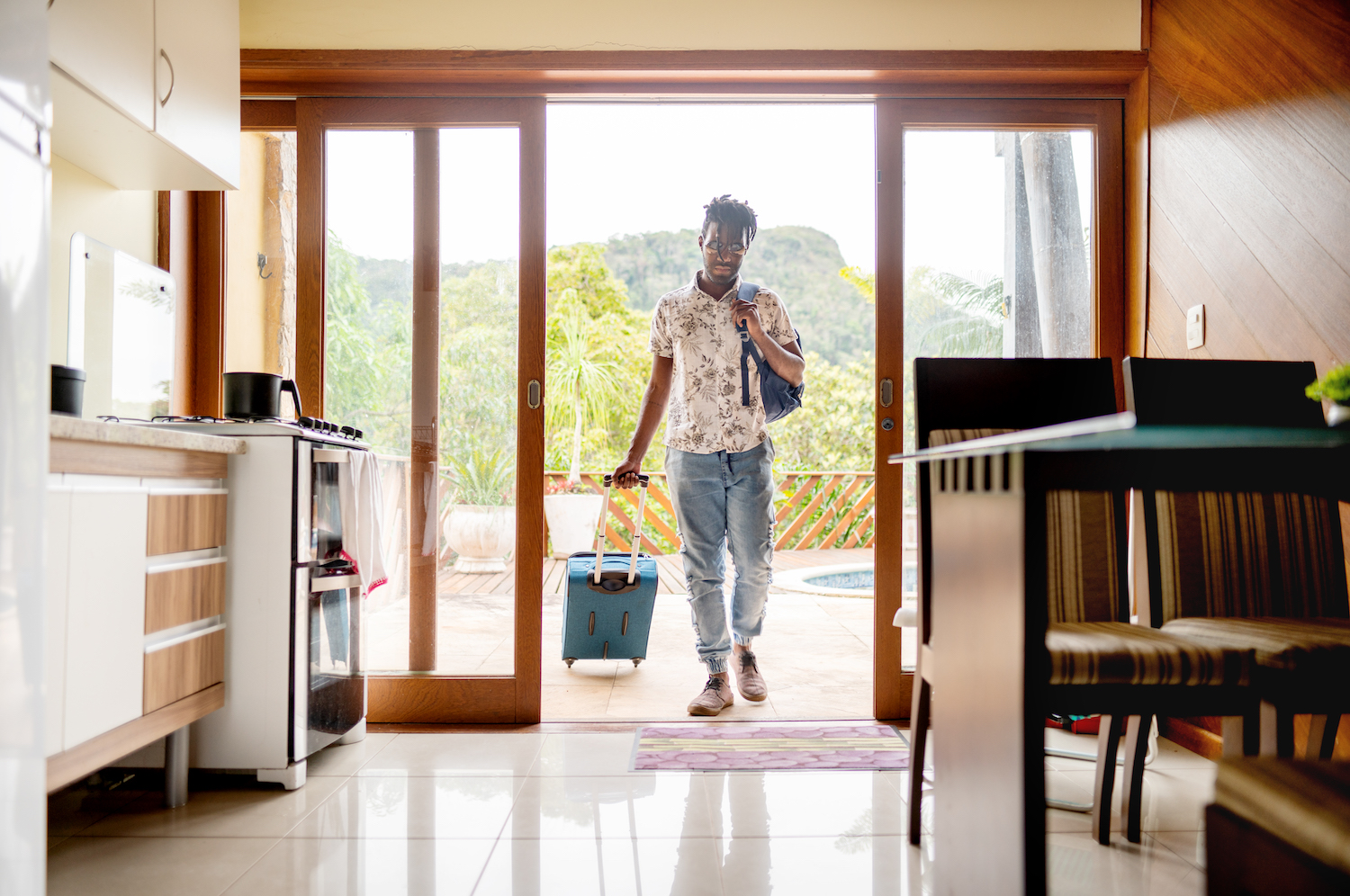 Young man walking through the patio doors of his rental accommodation pulling a wheeled suitcase