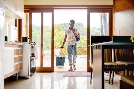 Young man walking through the patio doors of his rental accommodation pulling a wheeled suitcase