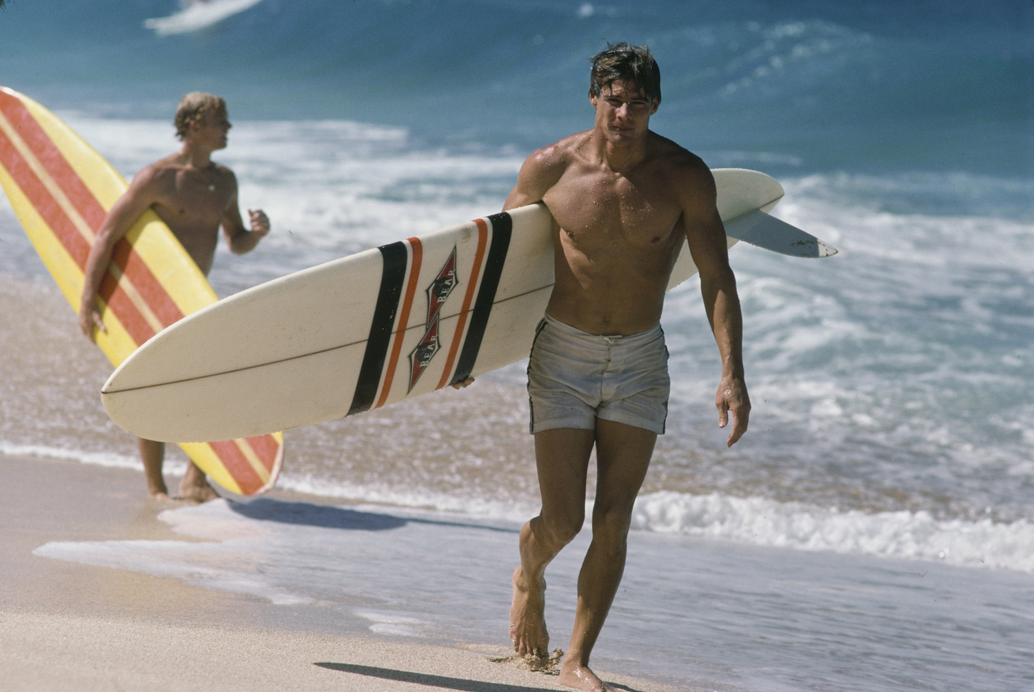 An actor playing a surfer, walking down the beach.