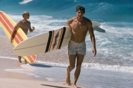 An actor playing a surfer, walking down the beach.