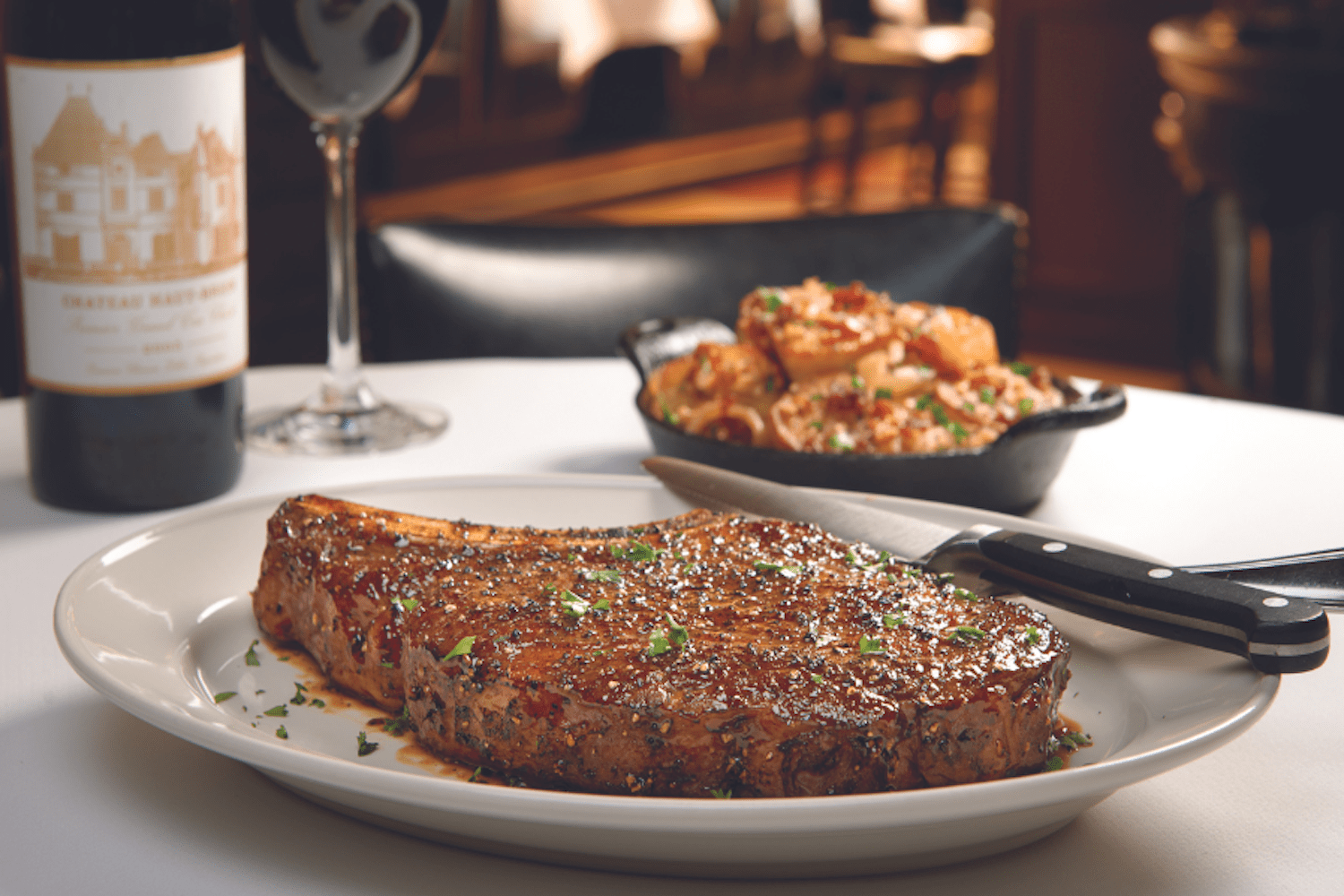 Steak on a plate on a white tablecloth behind wine and side dish.