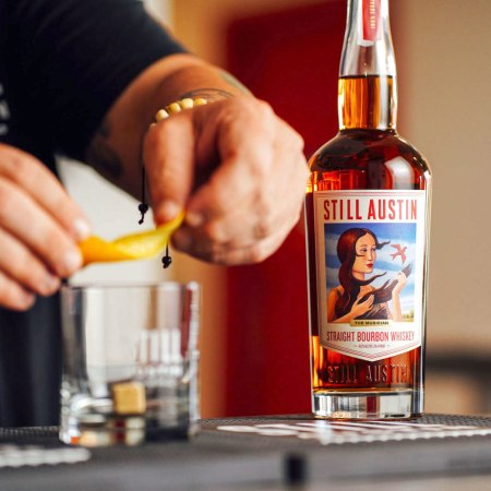 hands rimming a glass with lemon peel next to a bottle of whiskey