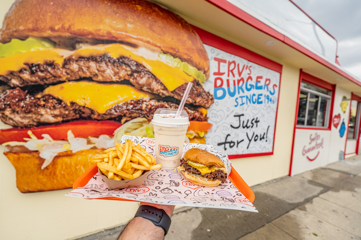 Burger, fries and a milkshake on a tray in front of a wall with a painted burger on it.