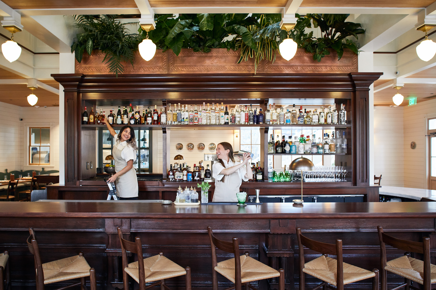 Two bartenders smiling behind the bar. One is shaking a cocktail shaker while the other reaches for a bottle on the shelf behind her.