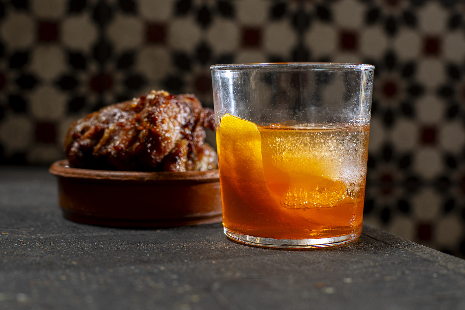 low-ball cocktail glass with lemon peel garnish in front of meat on a board.