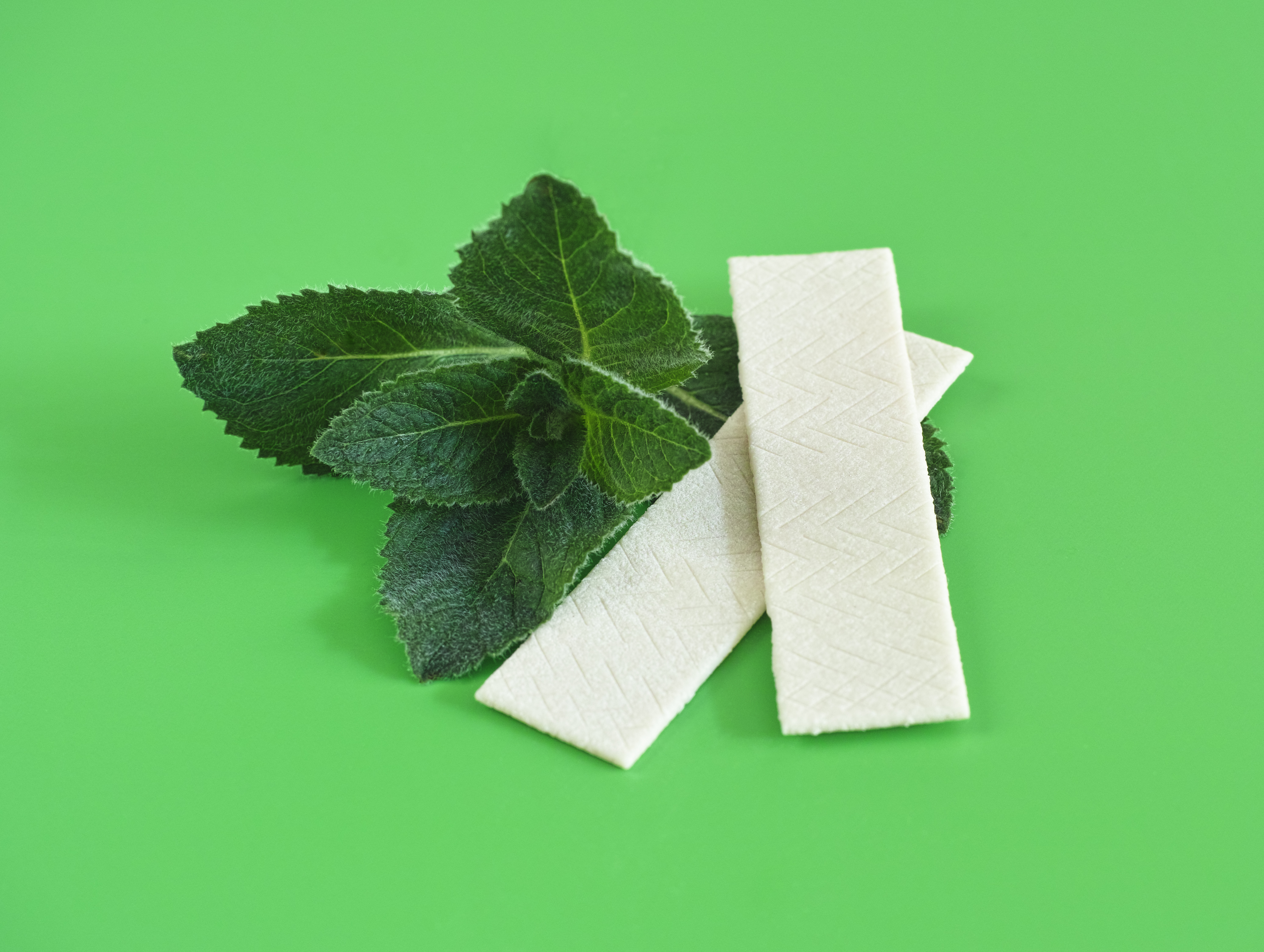 Two sticks of gum next to mint, against a green background.