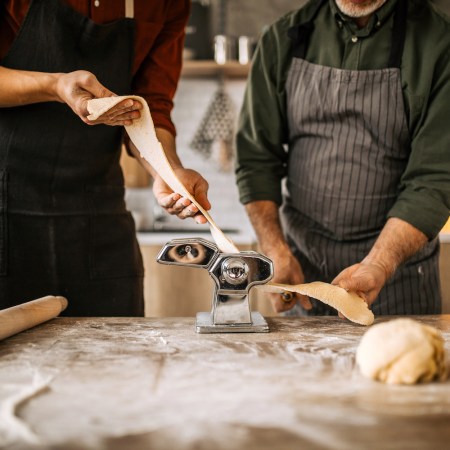Senior father and young adult son making homemade pasta, rolling out the pasta dough using the manual pasta maker in domestic kitchen