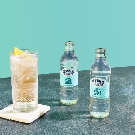 a whiskey highball next to Franklin & Sons club soda with a blue wall behind it