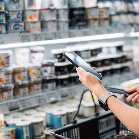 Cropped shot of young Asian woman holding shopping basket and checking her shopping list on smartphone while grocery shopping in a supermarket