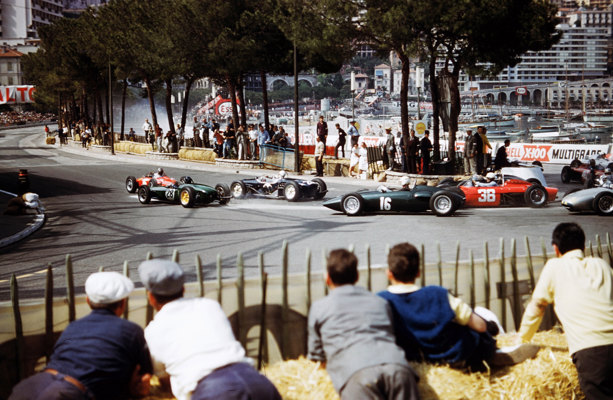 An old-school image of men crouched on a hill, watching racecars.