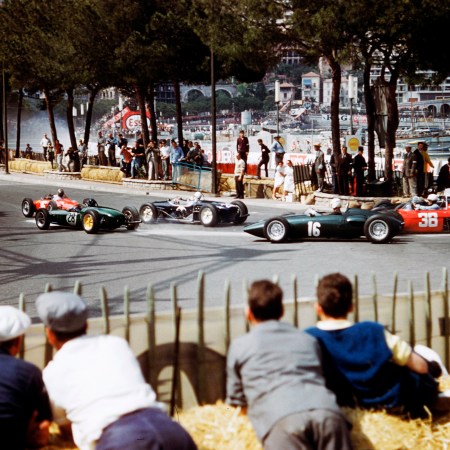 An old-school image of men crouched on a hill, watching racecars.