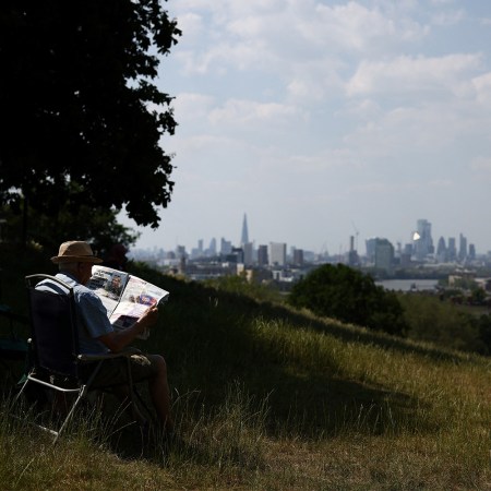A man reading a newspaper on a grassy hill, with a city in the distance.