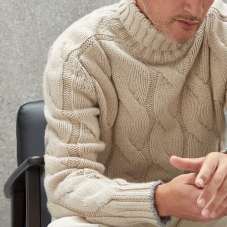 a model in a chunky turtleneck in a chair on a grey background