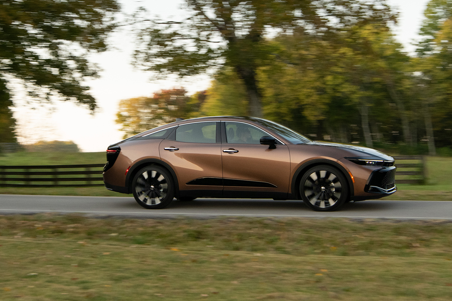 The new Toyota Crown sedan driving on a tree-lined road. In our review, we talk about the pros and cons of the hybrid car.