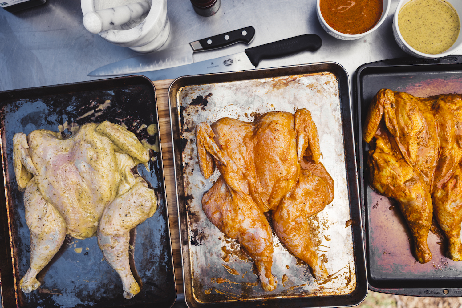 Three raw full chickens on a tray covered in different colored marinades