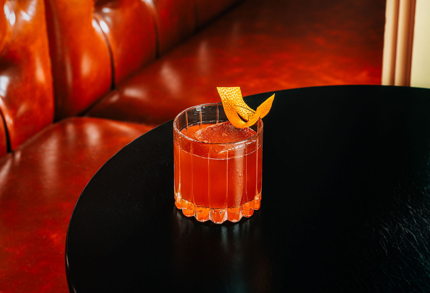 Lowball glass with orange-colored liquid and orange peel twist on a table in front of a red booth seat