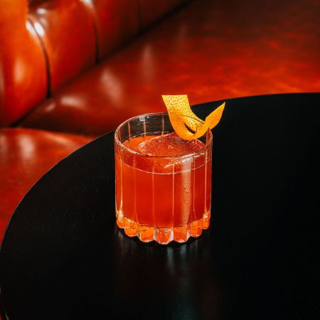 Lowball glass with orange-colored liquid and orange peel twist on a table in front of a red booth seat