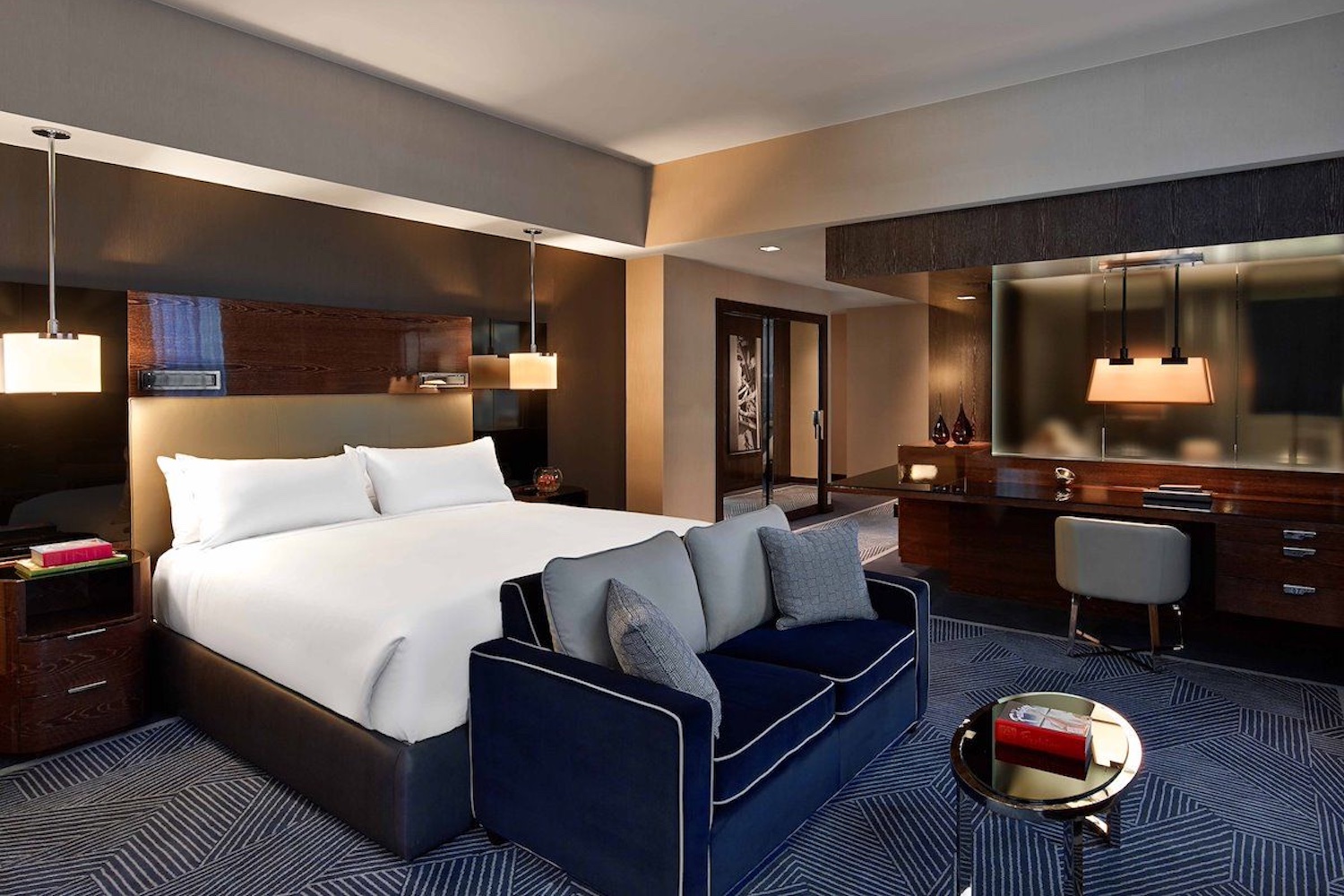 Bedroom with white bedding and dark-colored interior: gray carpets, a dark blue couch and dark wood accents