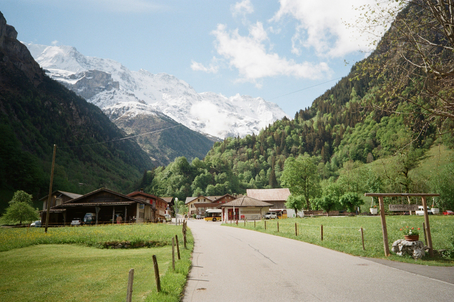 A road cuts through a scenic mountain valley with small Swiss huts not far away.