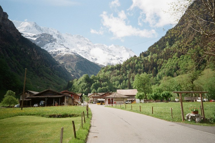 A road cuts through a scenic mountain valley with small Swiss huts not far away.