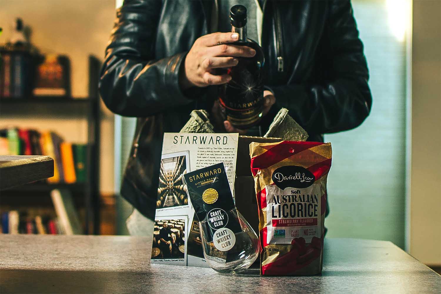 A person holding a bottle of Starward whisky with a basket of Australian food from the Craft Whisky Club in front of him