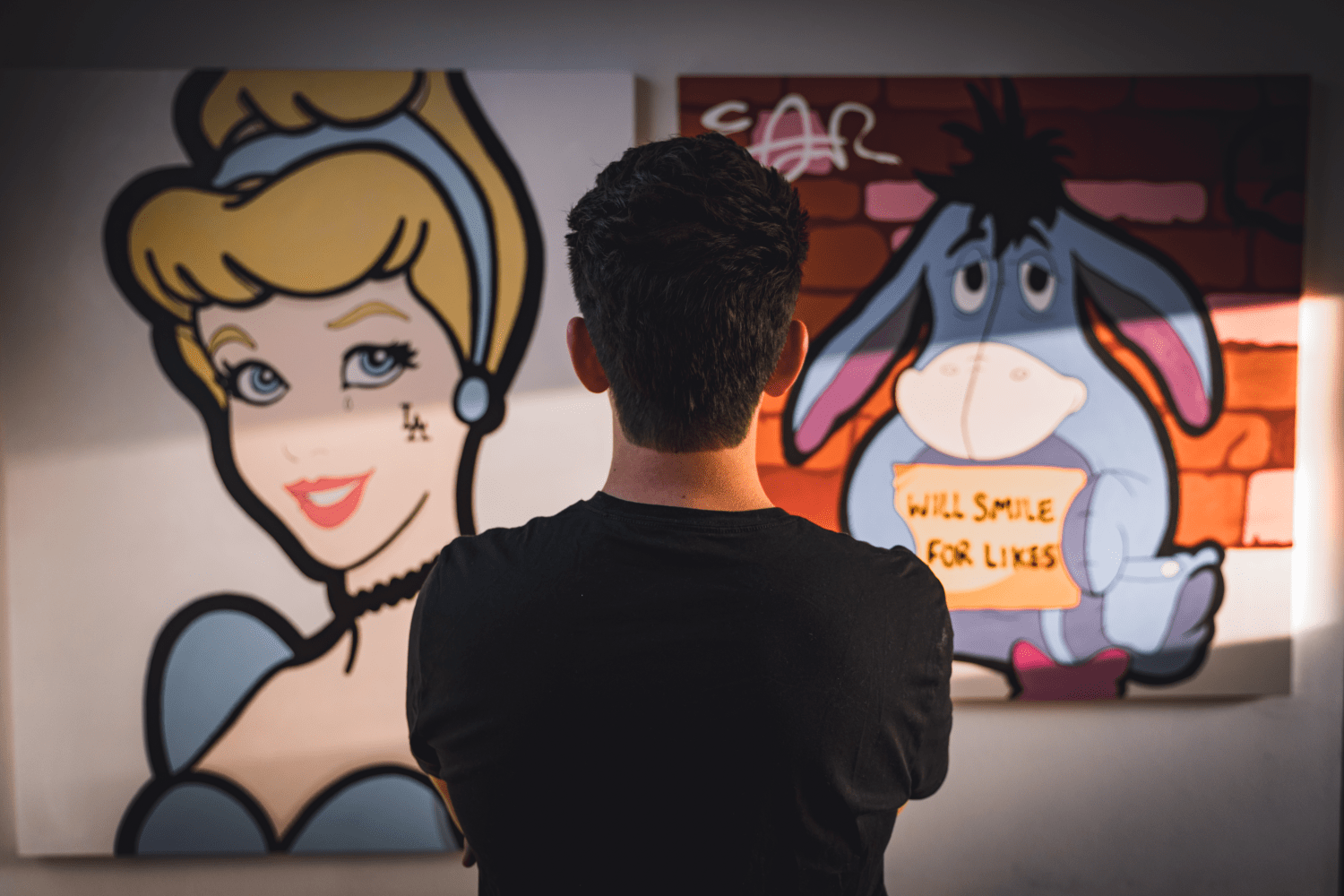 man looking at paintings of Cinderella with an LA face tattoo and eyore holding a sign that says will smile for likes