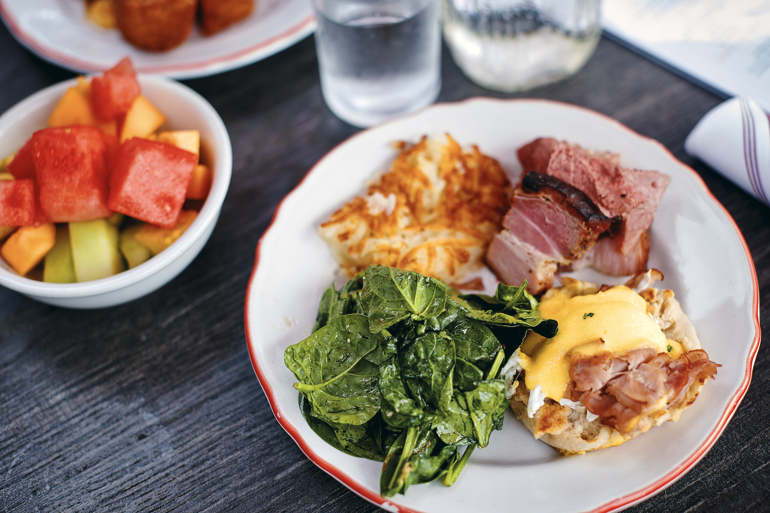 Plate with eggs benedict, greens, potatoes and bacon and a side cup of fruit