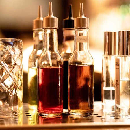 A glass and various cocktail bitters on a bar