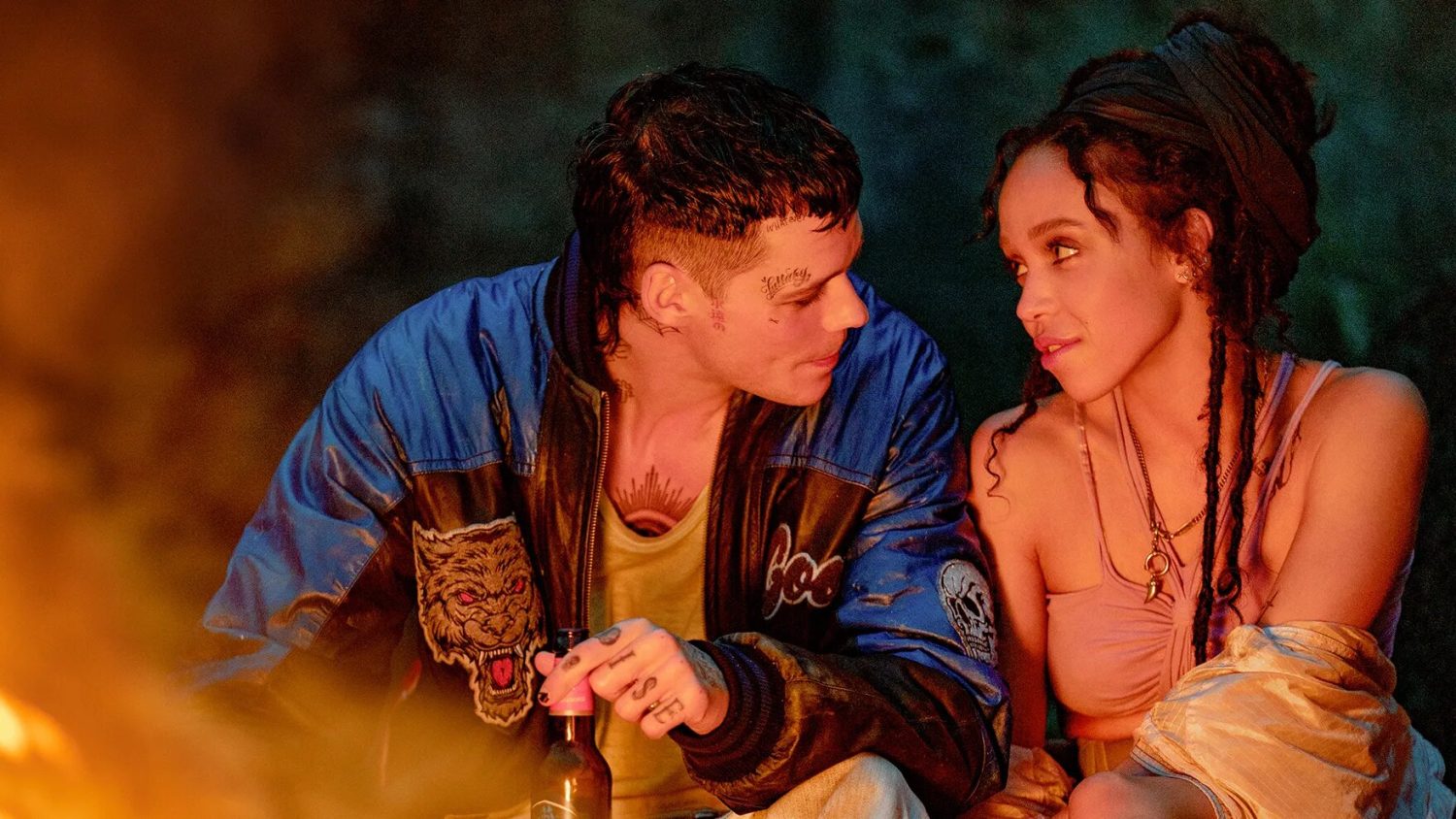Bill Skarsgard in a blue bomber jacket, with a mullet and face tattoos, looking at FKA Twigs in a dress
