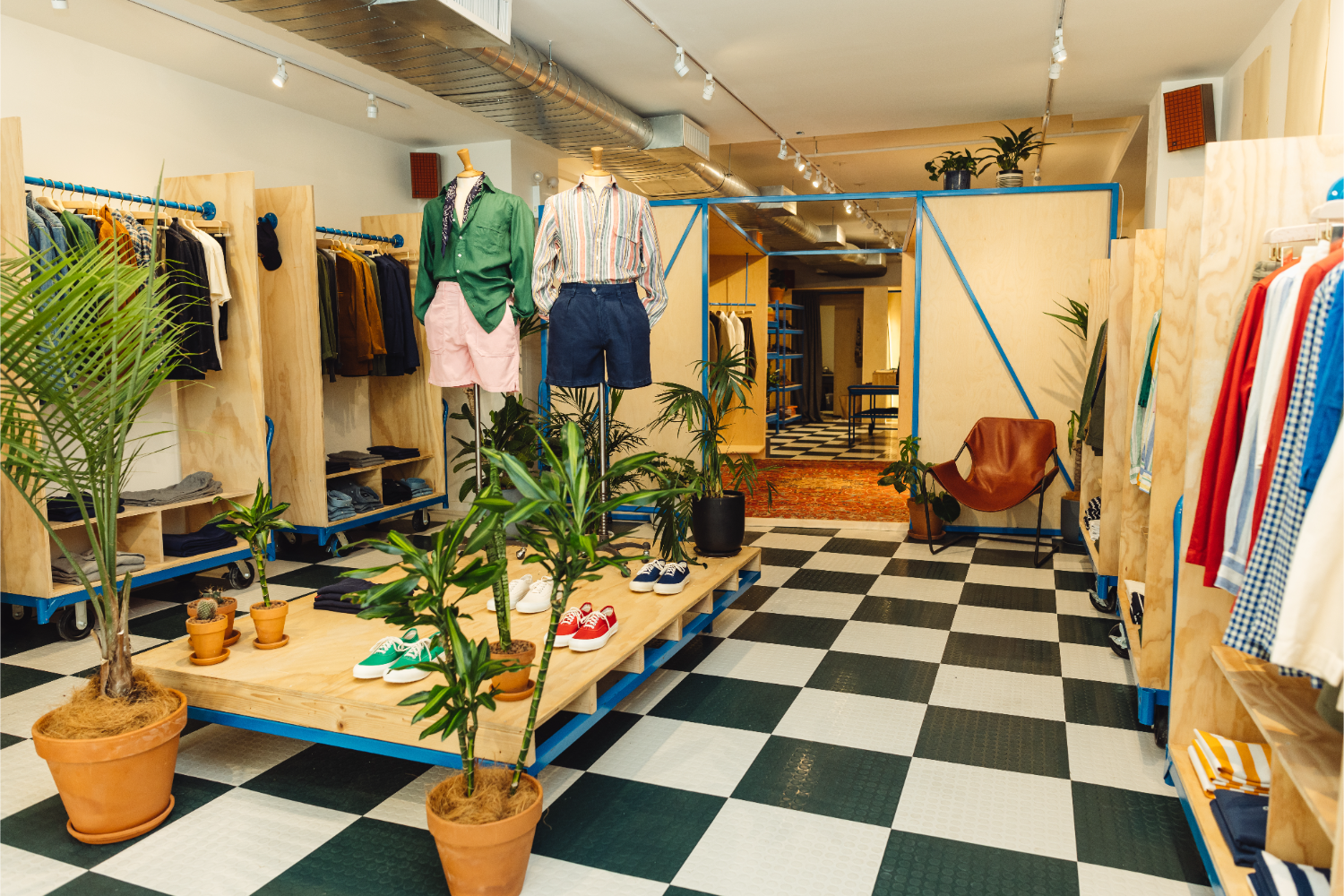 checkerboard floor, plants, mannequins with buttoned shirts and shorts, shoes on a wooden platform, clothes hanging