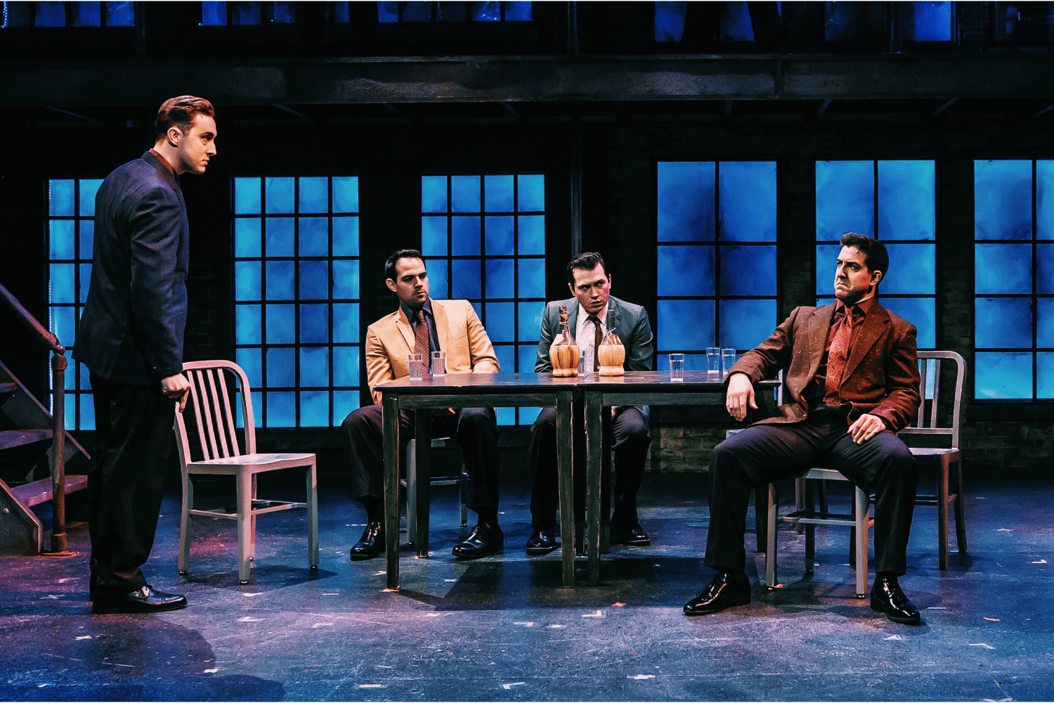 four male actors on stage in suits sitting in chairs, table