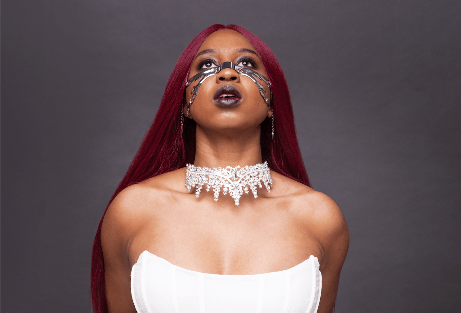woman with red hair staring up. silver face piece, black lipstick, white necklace and white shirt