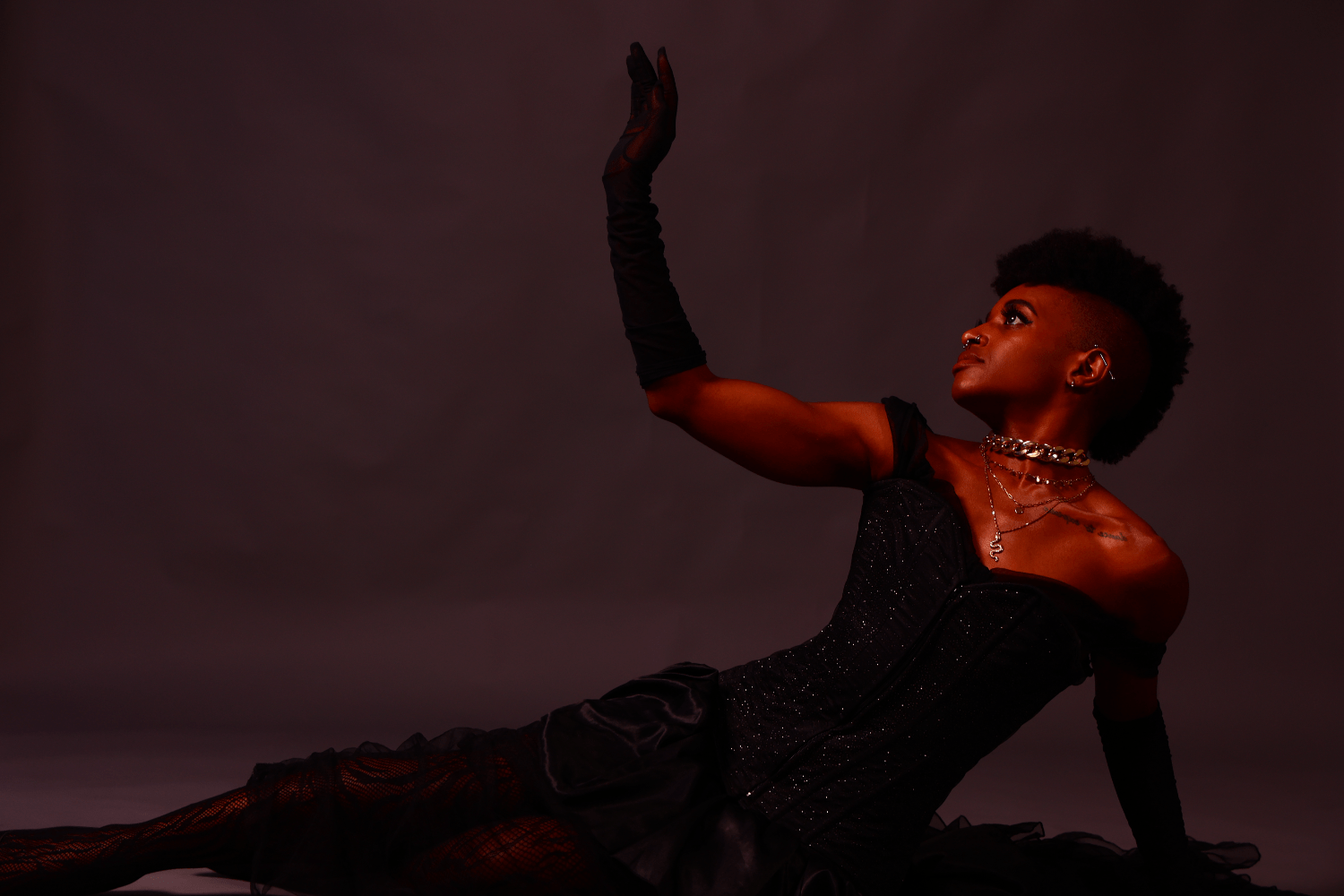 woman with piercings, silver jewelry, tattoo on collarbone and frohawk, in a black shimmery dress laying down sideways while looking up, arm out