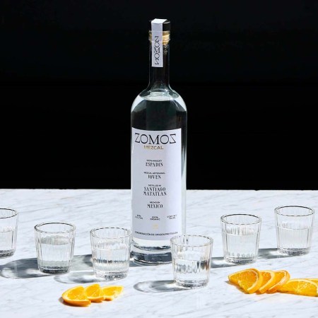 A bottle of Zomoz mezcal near orange slices and six shot glasses