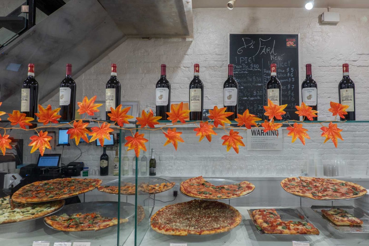 wine bottles lined up on top of glass display, orange decorative leaves, pizzas in the glass display