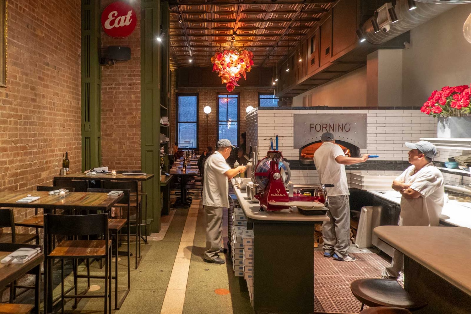 inside of pizzeria, two men in white clothes working putting pizza in brick oven, tables and lights