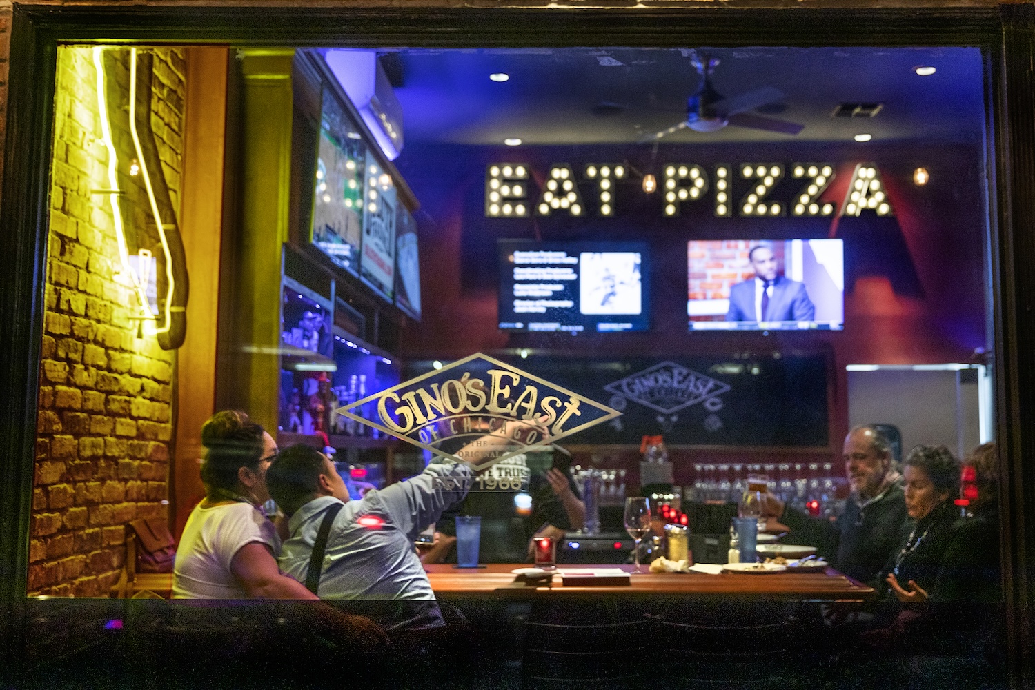 storefront window, eat pizza light up sign inside, couple sitting pointing, tvs
