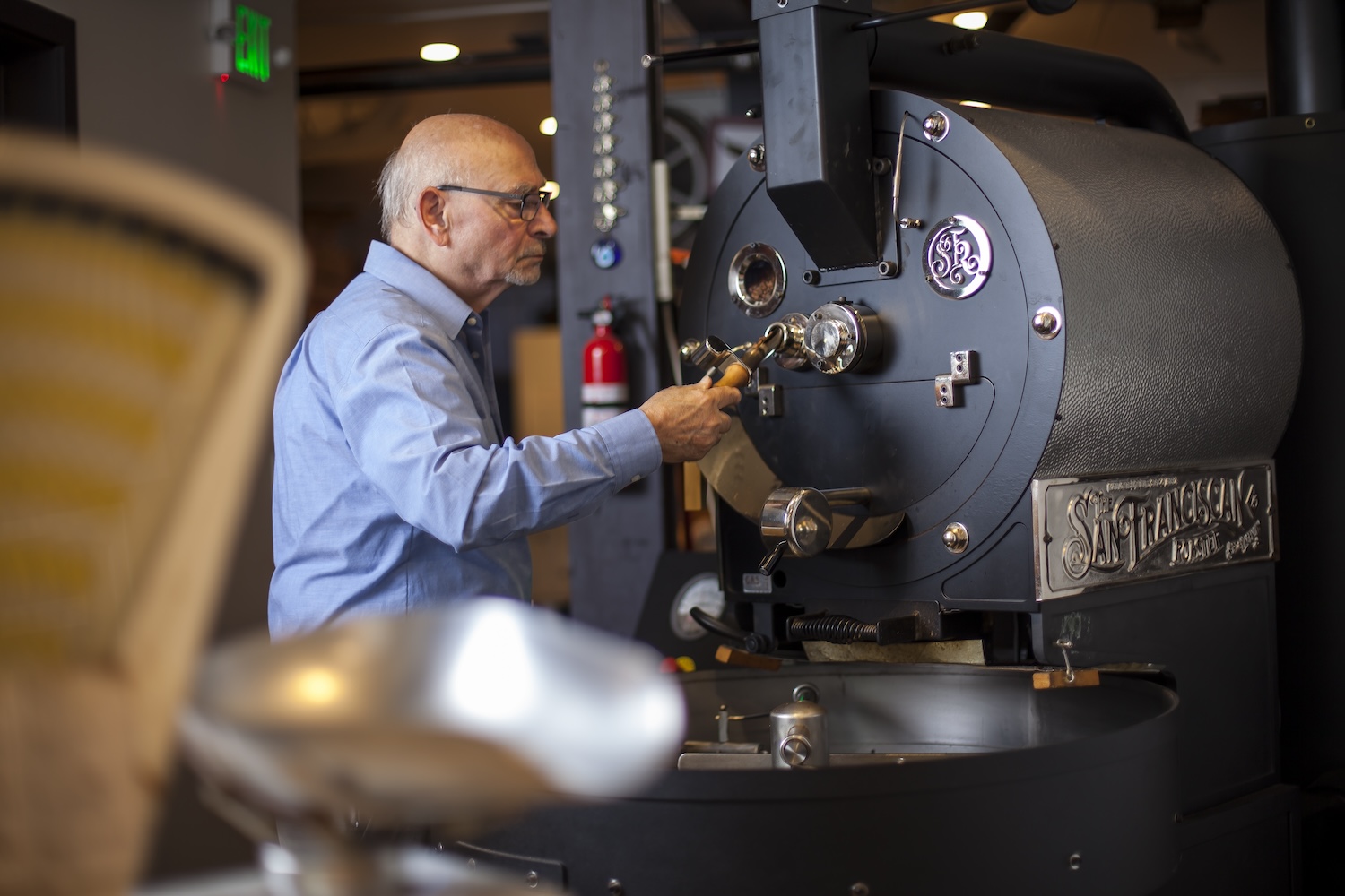 bald man in a blue buttoned-down shirt and glasses handling a coffee machine