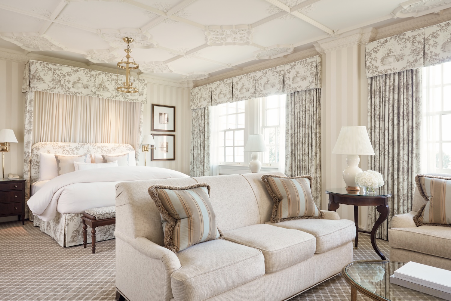 white room with white bed, beige couch with two pillows, window, table with lamp