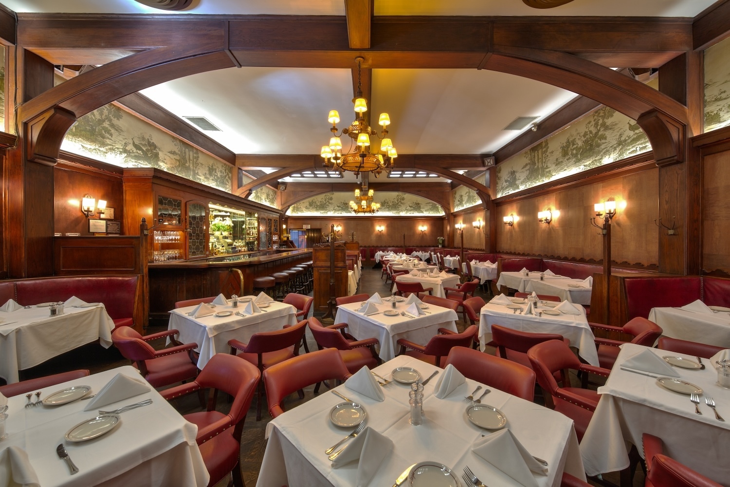 inside of restaurant with chandelier, lamps, tables with white tablecloth, glasses and utensils