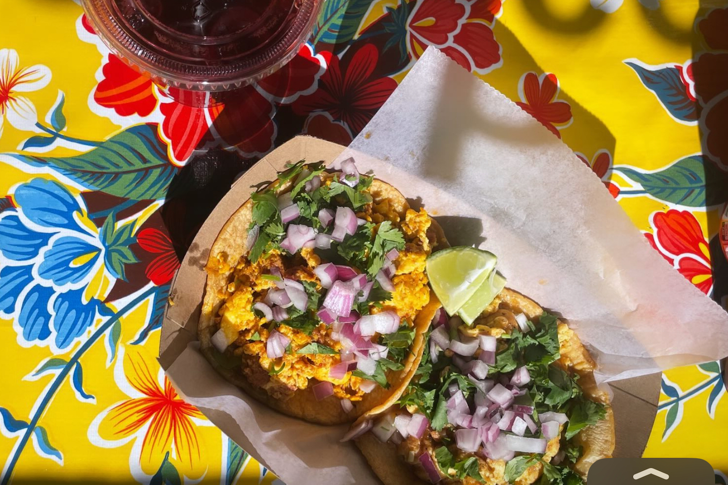 two tacos in a paper boat sitting on top of a yellow tablecloth with colorful flowers