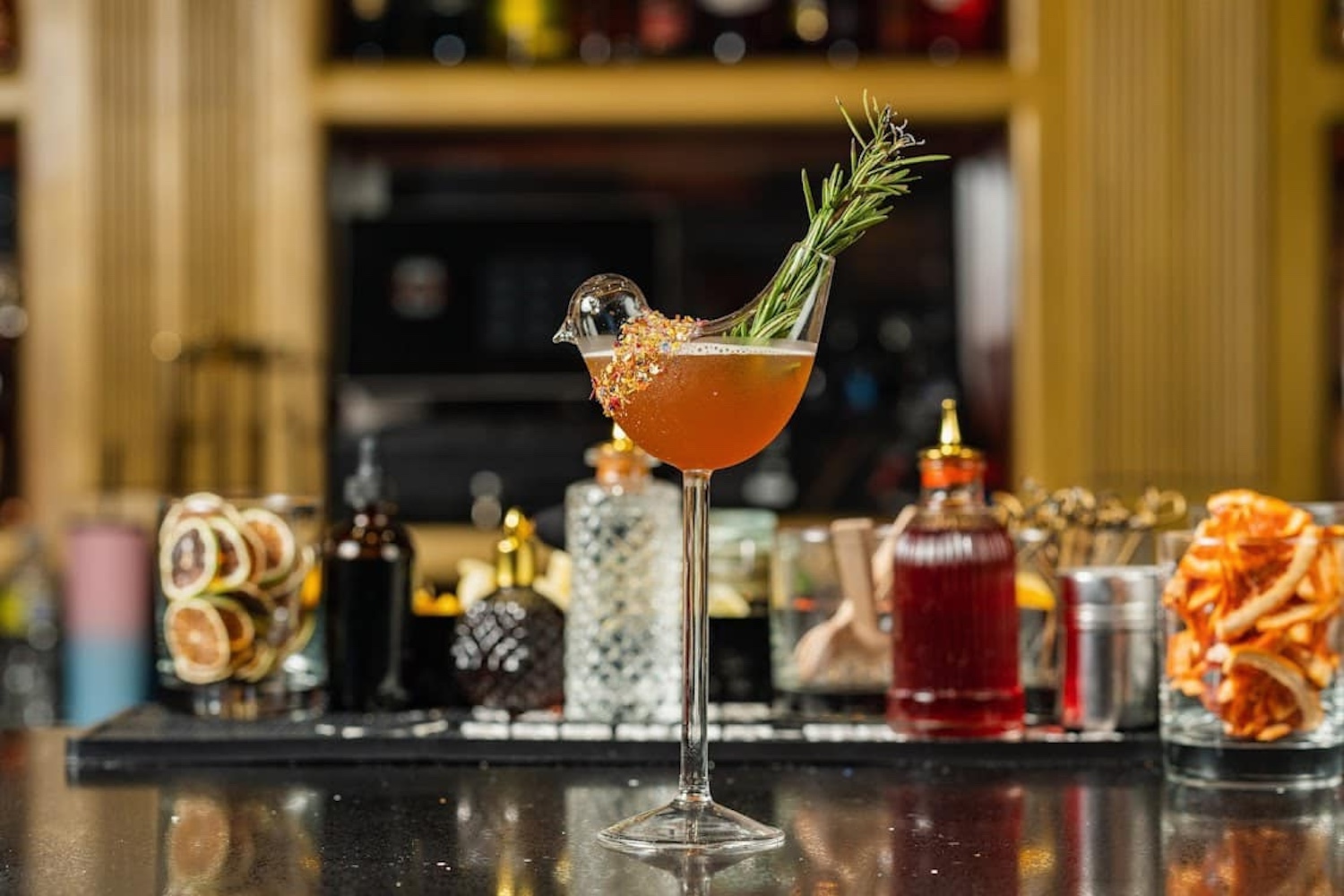 orange cocktail in a bird shaped glass with dried fruits and syrup in the background