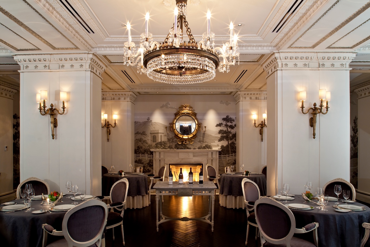 table in middle with bottles of champagne, fancy tables and seats with velvet cloth, gold mirror, columns with lamps on them, gold chandelier