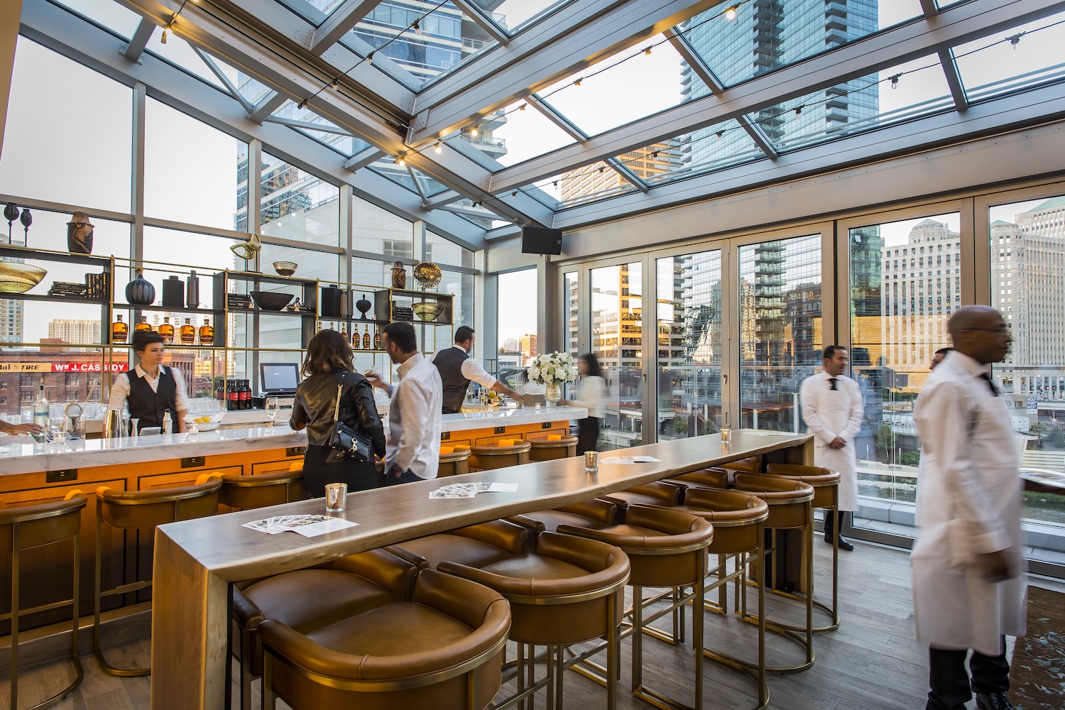 bar, glass window tent, long wooden table and brown chairs, servers in white uniforms, couple at the bar