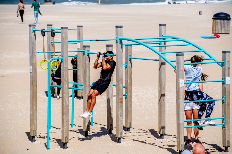 Two women, two kids and one man climbing and doing pull-ups on a jungle gym with blue bars on the sand