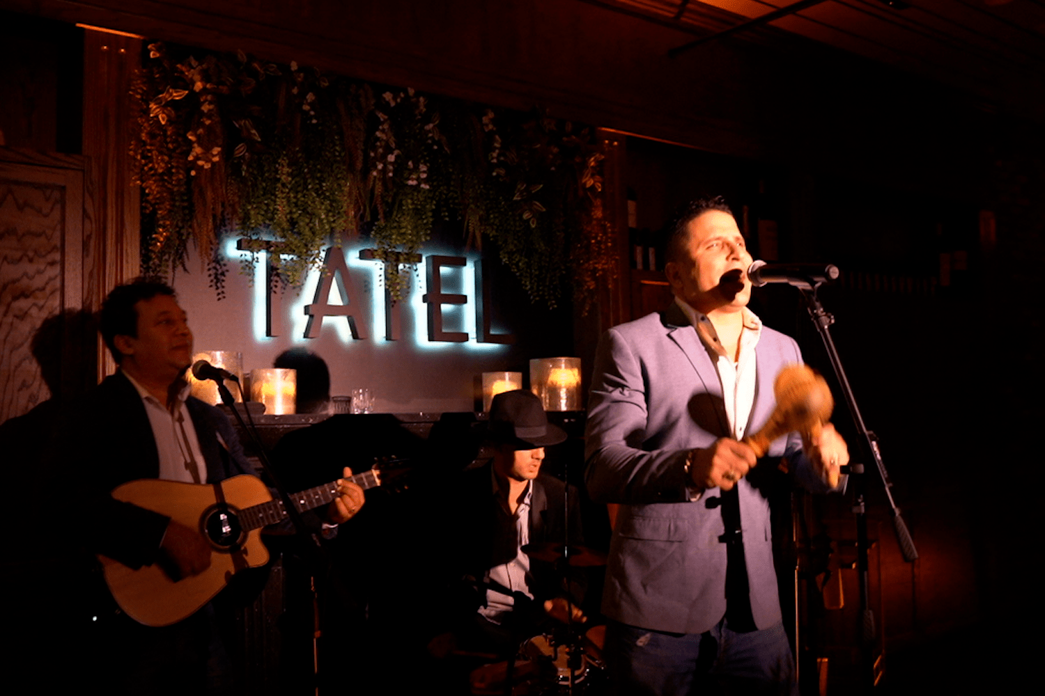 man shaking maracas and singing, man playing guitar behind him, light up sign in the background, flowers